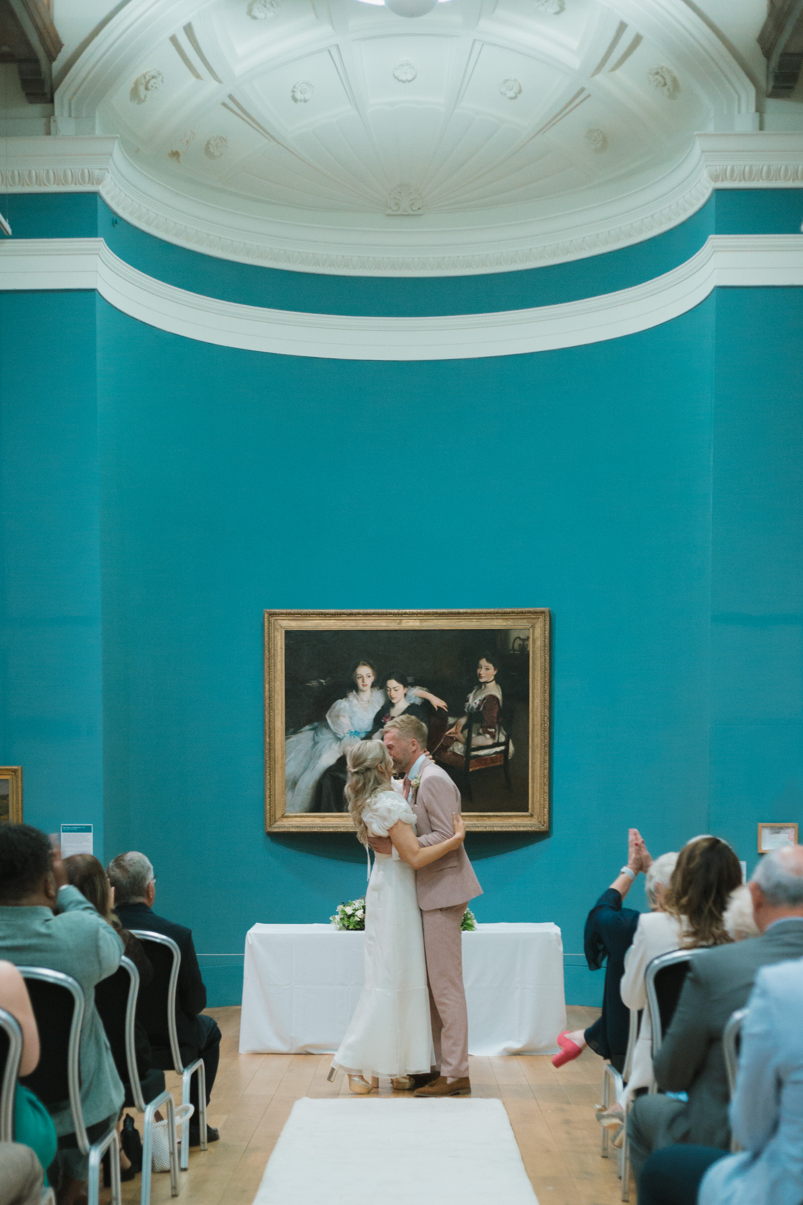 newly wed couple kissing at the alter in a museum with blue walls. Guests are clapping.